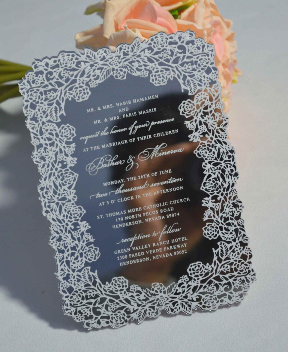 This image features a high-end silver mirror acrylic wedding invitation with an intricate white laser-cut lace border. The card is shown at an angle, leaning against a bouquet of soft peach roses on a white surface. The invitation features elegant white printed text with the details.
