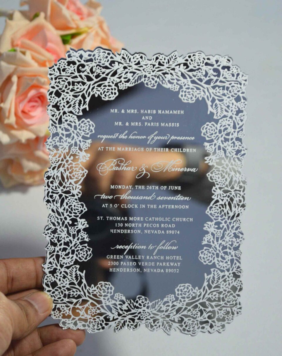 This image features a high-end silver mirror acrylic wedding invitation with an intricate white laser-cut lace border. The card is shown at an angle, leaning against a bouquet of soft peach roses on a white surface. The invitation features elegant white printed text with the details.