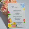 An elegant, rectangular wedding menu card is set against a background of draped, shimmering blush-pink satin and soft roses. The card features a delicate floral design with vibrant yellow, pink, and red flowers accenting the corners. The menu details are printed at the bottom. The overall aesthetic is romantic, colorful, and refined.