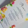 An elegant, rectangular wedding menu card is set against a background of draped, shimmering blush-pink satin and soft roses. The card features a delicate floral design with vibrant yellow, pink, and red flowers accenting the corners. The menu details are printed at the bottom. The overall aesthetic is romantic, colorful, and refined.