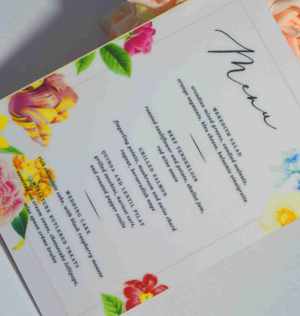 An elegant, rectangular wedding menu card is set against a background of draped, shimmering blush-pink satin and soft roses. The card features a delicate floral design with vibrant yellow, pink, and red flowers accenting the corners. The menu details are printed at the bottom. The overall aesthetic is romantic, colorful, and refined.