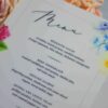 An elegant, rectangular wedding menu card is set against a background of draped, shimmering blush-pink satin and soft roses. The card features a delicate floral design with vibrant yellow, pink, and red flowers accenting the corners. The menu details are printed at the bottom. The overall aesthetic is romantic, colorful, and refined.