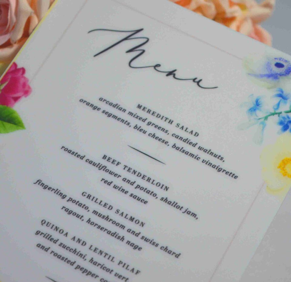 An elegant, rectangular wedding menu card is set against a background of draped, shimmering blush-pink satin and soft roses. The card features a delicate floral design with vibrant yellow, pink, and red flowers accenting the corners. The menu details are printed at the bottom. The overall aesthetic is romantic, colorful, and refined.