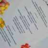 An elegant, rectangular wedding menu card is set against a background of draped, shimmering blush-pink satin and soft roses. The card features a delicate floral design with vibrant yellow, pink, and red flowers accenting the corners. The menu details are printed at the bottom. The overall aesthetic is romantic, colorful, and refined.