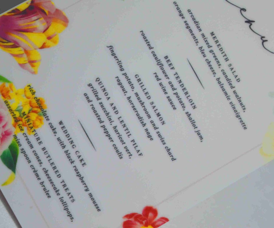 An elegant, rectangular wedding menu card is set against a background of draped, shimmering blush-pink satin and soft roses. The card features a delicate floral design with vibrant yellow, pink, and red flowers accenting the corners. The menu details are printed at the bottom. The overall aesthetic is romantic, colorful, and refined.