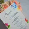 An elegant, rectangular wedding menu card is set against a background of draped, shimmering blush-pink satin and soft roses. The card features a delicate floral design with vibrant yellow, pink, and red flowers accenting the corners. The menu details are printed at the bottom. The overall aesthetic is romantic, colorful, and refined.