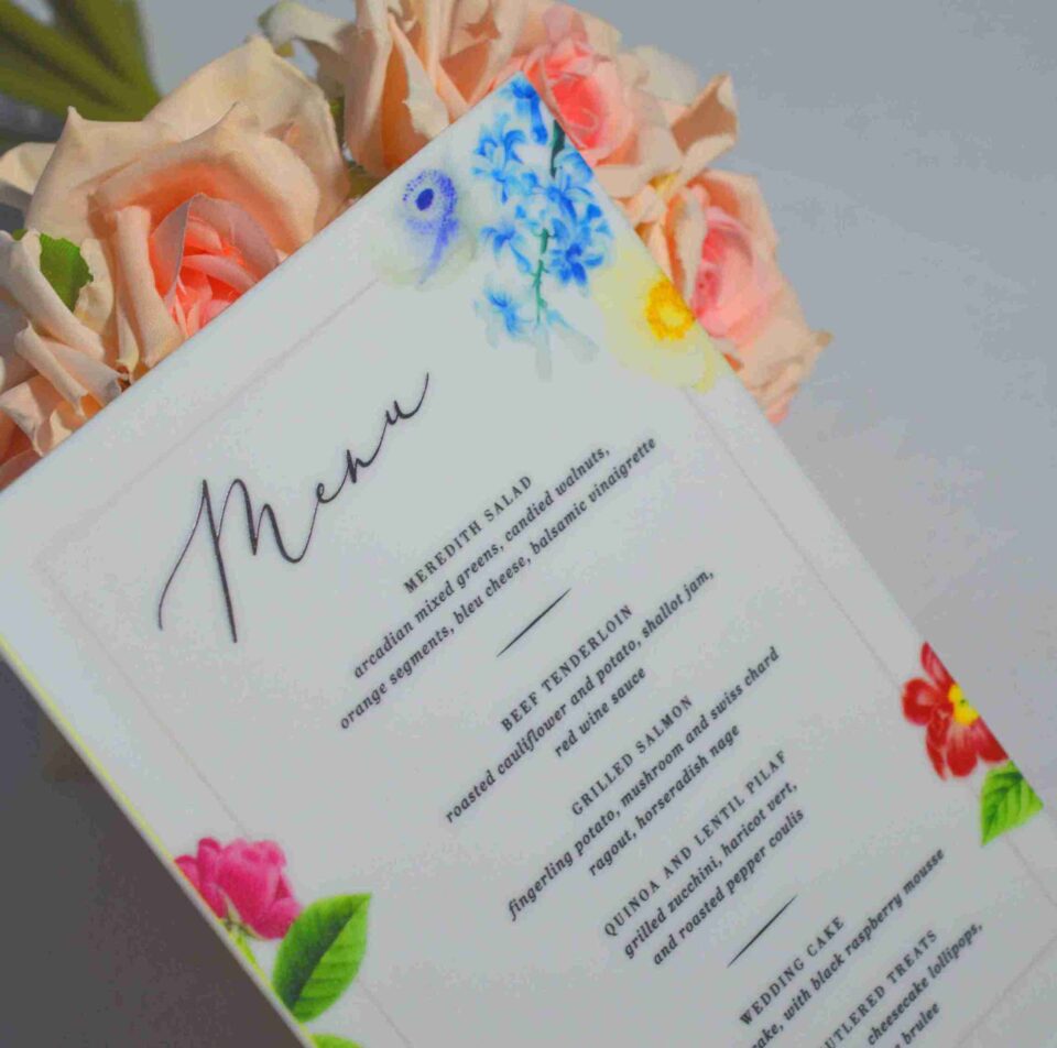 An elegant, rectangular wedding menu card is set against a background of draped, shimmering blush-pink satin and soft roses. The card features a delicate floral design with vibrant yellow, pink, and red flowers accenting the corners. The menu details are printed at the bottom. The overall aesthetic is romantic, colorful, and refined.