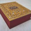 A high-angle, three-quarter view of a **luxurious burgundy invitation box** with an ornate, multi-layered lid. The box features: * A **laser-cut gold wooden overlay** on the lid, showcasing an intricate floral and scrollwork pattern. * A central **maroon plaque** featuring the custom monogram "R & F" etched in gold. * A border of **sparkling clear crystals** lining the inner edge of the gold frame. * The deep burgundy velvet sides provide a rich, tactile contrast to the metallic gold top. * The box is displayed on a neutral, textured light-colored surface, emphasizing its elegant and substantial design.