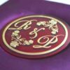 A low-angle shot of a square, vibrant purple silk invitation box resting on a marbled white surface. The padded lid features a central circular acrylic plaque in deep burgundy with a gold-foil border. The plaque is decorated with an elegant gold monogram "R & L" intertwined with delicate floral illustrations.