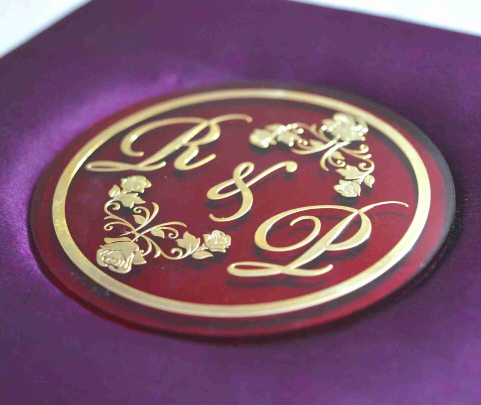 A low-angle shot of a square, vibrant purple silk invitation box resting on a marbled white surface. The padded lid features a central circular acrylic plaque in deep burgundy with a gold-foil border. The plaque is decorated with an elegant gold monogram "R & L" intertwined with delicate floral illustrations.