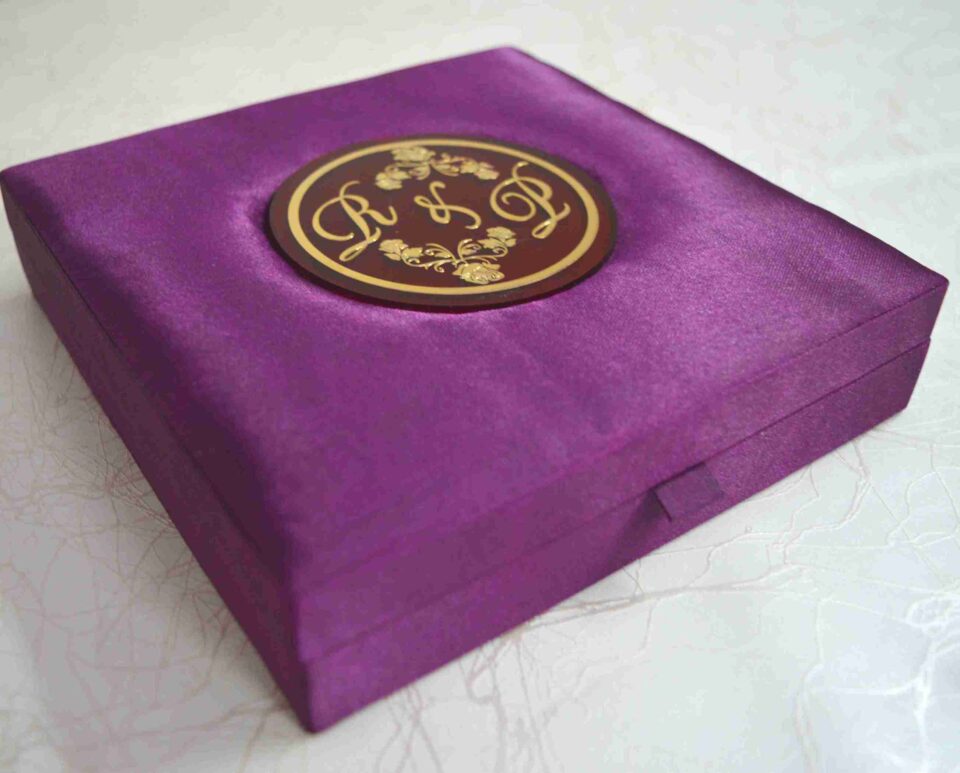 A low-angle shot of a square, vibrant purple silk invitation box resting on a marbled white surface. The padded lid features a central circular acrylic plaque in deep burgundy with a gold-foil border. The plaque is decorated with an elegant gold monogram "R & L" intertwined with delicate floral illustrations.
