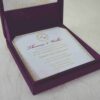 An open, square purple silk invitation box resting on a white marbled surface. Nested inside the box is a formal white wedding invitation for Thomas and Wallis, featuring a gold-scrolled border and a central gold monogram. In the foreground, a matching white RSVP card is displayed, decorated with gold floral scrollwork and an "RSVP" heading in elegant script. Another card with the names Victoria and Patric is partially visible on the left.