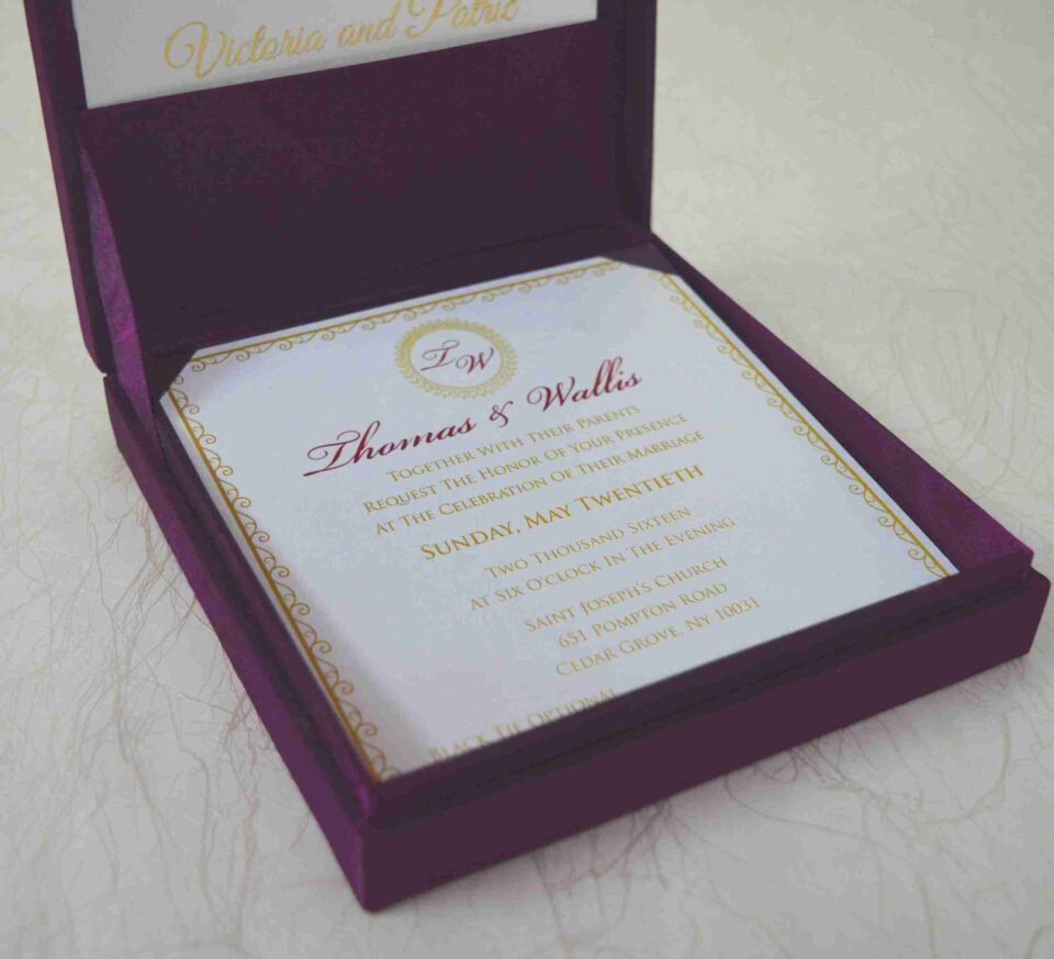 An open, square purple silk invitation box resting on a white marbled surface. Nested inside the box is a formal white wedding invitation for Thomas and Wallis, featuring a gold-scrolled border and a central gold monogram. In the foreground, a matching white RSVP card is displayed, decorated with gold floral scrollwork and an "RSVP" heading in elegant script. Another card with the names Victoria and Patric is partially visible on the left.