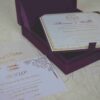 An open, square purple silk invitation box resting on a white marbled surface. Nested inside the box is a formal white wedding invitation for Thomas and Wallis, featuring a gold-scrolled border and a central gold monogram. In the foreground, a matching white RSVP card is displayed, decorated with gold floral scrollwork and an "RSVP" heading in elegant script. Another card with the names Victoria and Patric is partially visible on the left.
