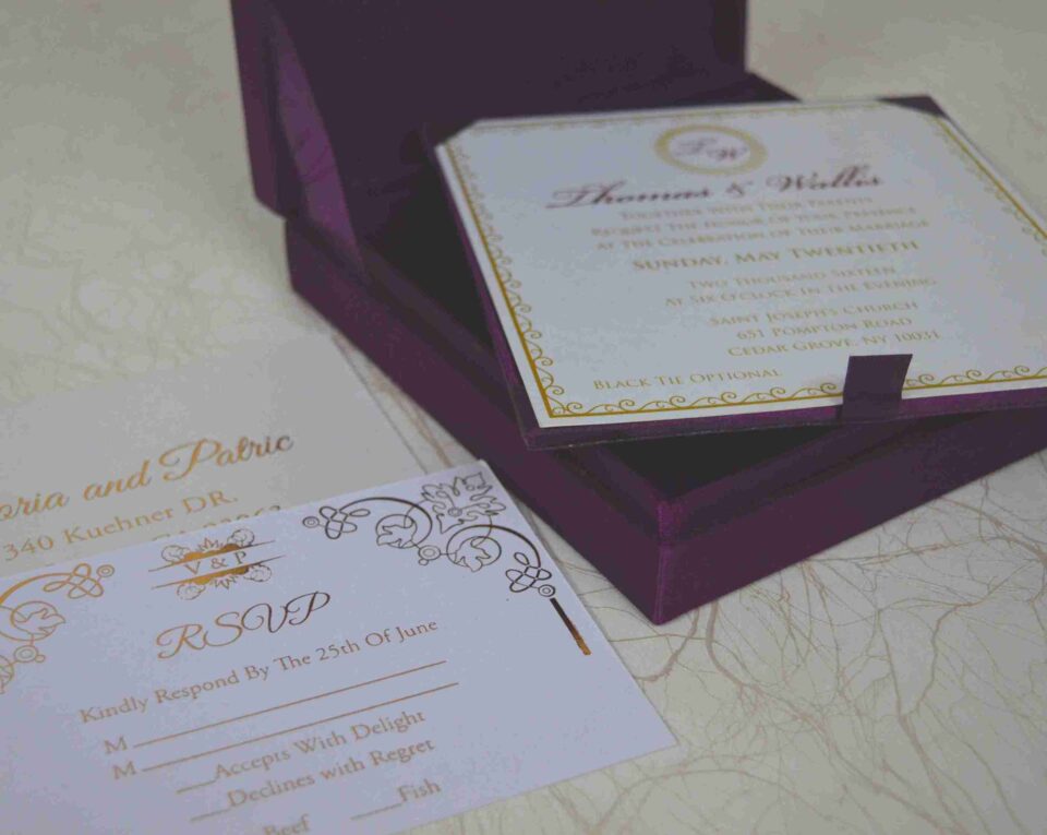 An open, square purple silk invitation box resting on a white marbled surface. Nested inside the box is a formal white wedding invitation for Thomas and Wallis, featuring a gold-scrolled border and a central gold monogram. In the foreground, a matching white RSVP card is displayed, decorated with gold floral scrollwork and an "RSVP" heading in elegant script. Another card with the names Victoria and Patric is partially visible on the left.