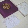 An open, square purple silk invitation box resting on a white marbled surface. Nested inside the box is a formal white wedding invitation for Thomas and Wallis, featuring a gold-scrolled border and a central gold monogram. In the foreground, a matching white RSVP card is displayed, decorated with gold floral scrollwork and an "RSVP" heading in elegant script. Another card with the names Victoria and Patric is partially visible on the left.