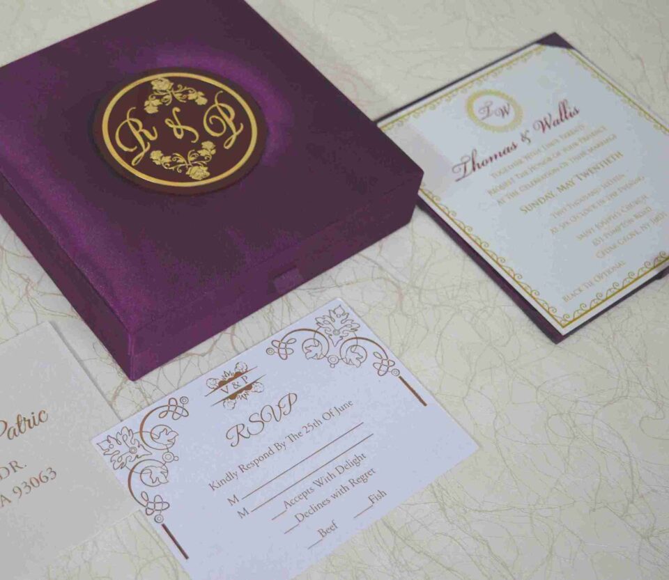 An open, square purple silk invitation box resting on a white marbled surface. Nested inside the box is a formal white wedding invitation for Thomas and Wallis, featuring a gold-scrolled border and a central gold monogram. In the foreground, a matching white RSVP card is displayed, decorated with gold floral scrollwork and an "RSVP" heading in elegant script. Another card with the names Victoria and Patric is partially visible on the left.
