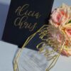An elegant wedding invitation set featuring a black rectangular box and a geometric acrylic invitation. The black box on the left has written in gold cursive foil. To the right, a clear, geometric-shaped acrylic invitation rests against a bouquet of peach-colored roses. The acrylic piece also features the name in gold mirror acrylic script at the top, with the wedding details printed in smaller gold text below. The entire set is presented on a light blue surface.