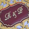 A high-angle, three-quarter view of a **luxurious burgundy invitation box** with an ornate, multi-layered lid. The box features: * A **laser-cut gold wooden overlay** on the lid, showcasing an intricate floral and scrollwork pattern. * A central **maroon plaque** featuring the custom monogram "R & F" etched in gold. * A border of **sparkling clear crystals** lining the inner edge of the gold frame. * The deep burgundy velvet sides provide a rich, tactile contrast to the metallic gold top. * The box is displayed on a neutral, textured light-colored surface, emphasizing its elegant and substantial design.