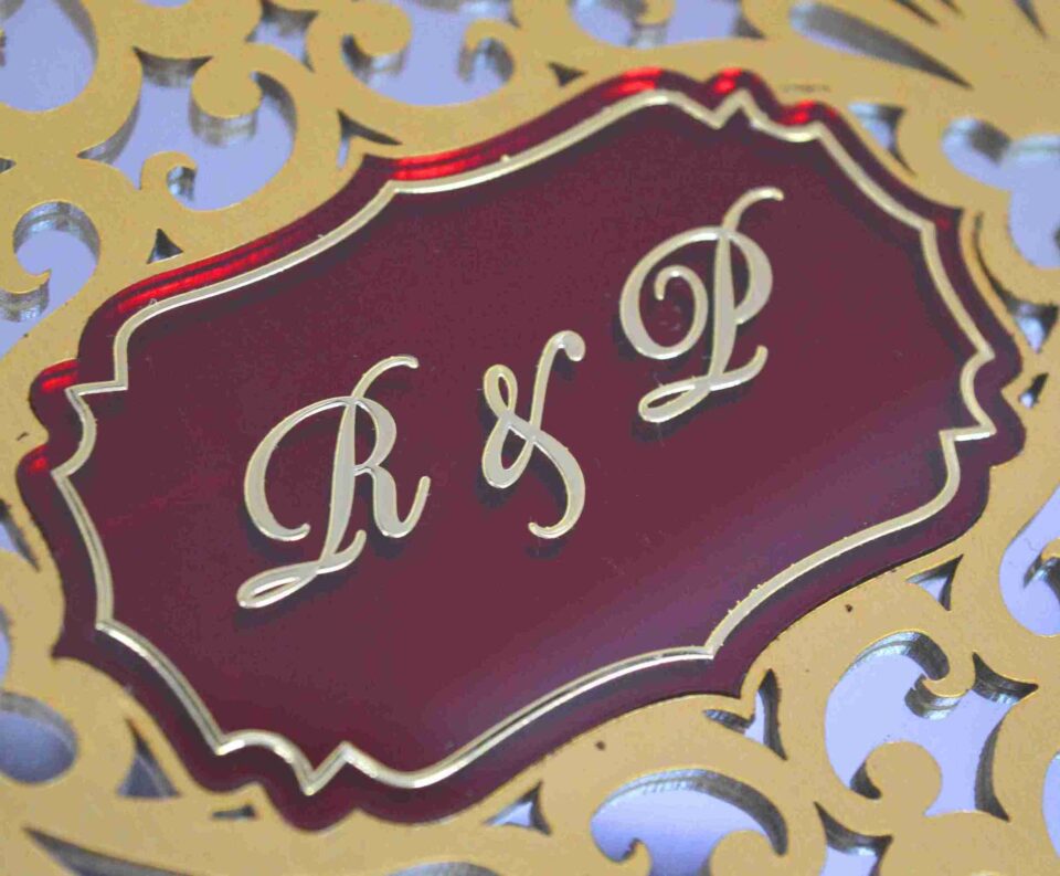 A high-angle, three-quarter view of a **luxurious burgundy invitation box** with an ornate, multi-layered lid. The box features: * A **laser-cut gold wooden overlay** on the lid, showcasing an intricate floral and scrollwork pattern. * A central **maroon plaque** featuring the custom monogram "R & F" etched in gold. * A border of **sparkling clear crystals** lining the inner edge of the gold frame. * The deep burgundy velvet sides provide a rich, tactile contrast to the metallic gold top. * The box is displayed on a neutral, textured light-colored surface, emphasizing its elegant and substantial design.