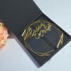 An elegant wedding invitation set featuring a black rectangular box and a geometric acrylic invitation. The black box on the left has written in gold cursive foil. To the right, a clear, geometric-shaped acrylic invitation rests against a bouquet of peach-colored roses. The acrylic piece also features the name in gold mirror acrylic script at the top, with the wedding details printed in smaller gold text below. The entire set is presented on a light blue surface.