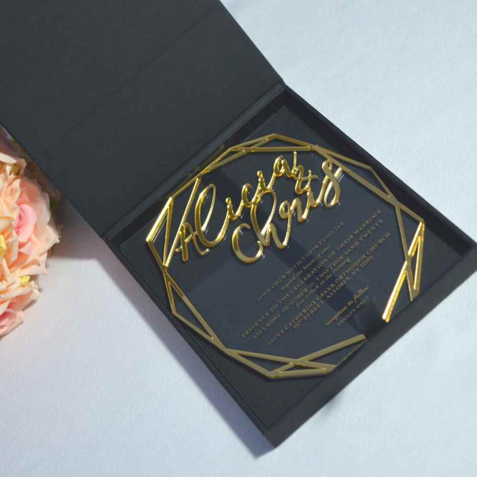 An elegant wedding invitation set featuring a black rectangular box and a geometric acrylic invitation. The black box on the left has written in gold cursive foil. To the right, a clear, geometric-shaped acrylic invitation rests against a bouquet of peach-colored roses. The acrylic piece also features the name in gold mirror acrylic script at the top, with the wedding details printed in smaller gold text below. The entire set is presented on a light blue surface.