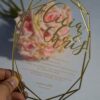 A close-up of a hand holding a unique, geometric-shaped clear acrylic wedding invitation.The card features a thin gold-mirrored border following its multi-faceted, decagon-like silhouette. The design includes: The name prominently etched in a large, elegant gold script at the top. Formal event details printed in gold serif font below, announcing a ceremony date, location Church The background is softly blurred, showing a bouquet of pink and peach roses, which enhances the luxurious and modern aesthetic of the invitation.