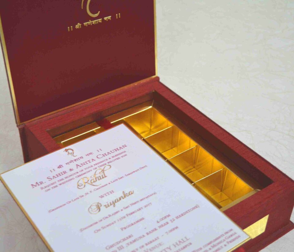 An open, **luxurious maroon wedding invitation box** featuring a multi-functional design with a built-in sweets compartment. The interior includes: * **The Invitation:** A white card printed with elegant red and gold typography for the wedding of "Rahul with Priyanka," featuring traditional Hindu motifs and "Shree Ganeshay Namah" script. * **Sweets Compartment:** To the right of the invitation, a **gold-foiled grid** provides individual sections for chocolates or traditional sweets. * **Inner Lid:** The maroon lid is accented with a thin gold border and a central gold-foiled religious inscription. * The box has a deep, textured wood-grain or fabric finish, giving it a high-end and traditional look. * The entire piece is set against a neutral, light-colored background, emphasizing the rich color palette of maroon and gold.