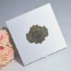 A top-down, slightly angled view of a square, **white shimmering silk invitation box. In the center of the lid is a large, **ornate gold metal plaque** with an embossed oval crest containing stylized initials. A bouquet of **soft peach and pink roses** is visible in the lower-left corner, and the box sits on a smooth, light grey surface.