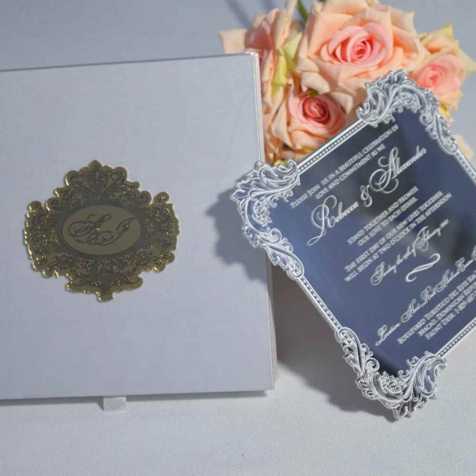 An open, **white silk invitation folio** featuring a large, ornate gold monogram plaque on the left panel. Leaning against the right side is a **clear acrylic wedding invitation** with intricate silver-etched borders and white script lettering for the names "**Rebecca & Alexander**." The invitation details a ceremony at the Hard Rock Hotel and Casino. In the background, soft peach-colored roses add a delicate floral accent against a neutral grey surface.