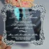 A close-up of a square clear acrylic wedding invitation held by a hand. The invitation features an elaborate, silver-etched floral scrollwork border** and elegant white text. The names "Rebecca & Alexander" are written in a large, graceful script. The invitation details a ceremony on Sunday, the 14th of February, 2021, at the **Hard Rock Hotel and Casino Punta Cana. The transparent card reflects a dark background, and a soft peach rose is blurred in the top background.