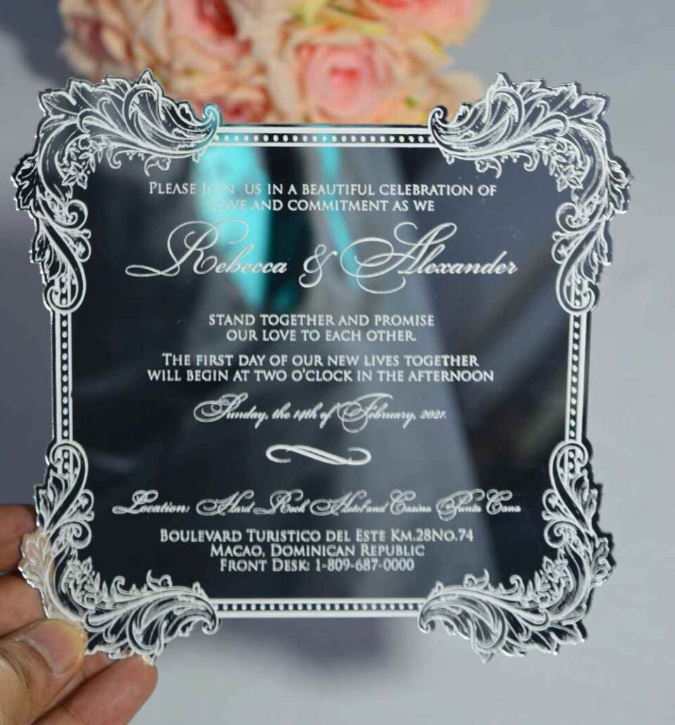 A close-up of a square clear acrylic wedding invitation held by a hand. The invitation features an elaborate, silver-etched floral scrollwork border** and elegant white text. The names "Rebecca & Alexander" are written in a large, graceful script. The invitation details a ceremony on Sunday, the 14th of February, 2021, at the **Hard Rock Hotel and Casino Punta Cana. The transparent card reflects a dark background, and a soft peach rose is blurred in the top background.