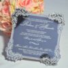A square wedding invitation made of a highly reflective silver mirror acrylic material. The invitation features an ornate, white baroque-style floral border in each of the four corners and along the scalloped edges The mirror surface is so reflective that the photographer holding a camera is visible in the center of the card. A bouquet of peach-colored roses is partially visible in the blurred background on the left.