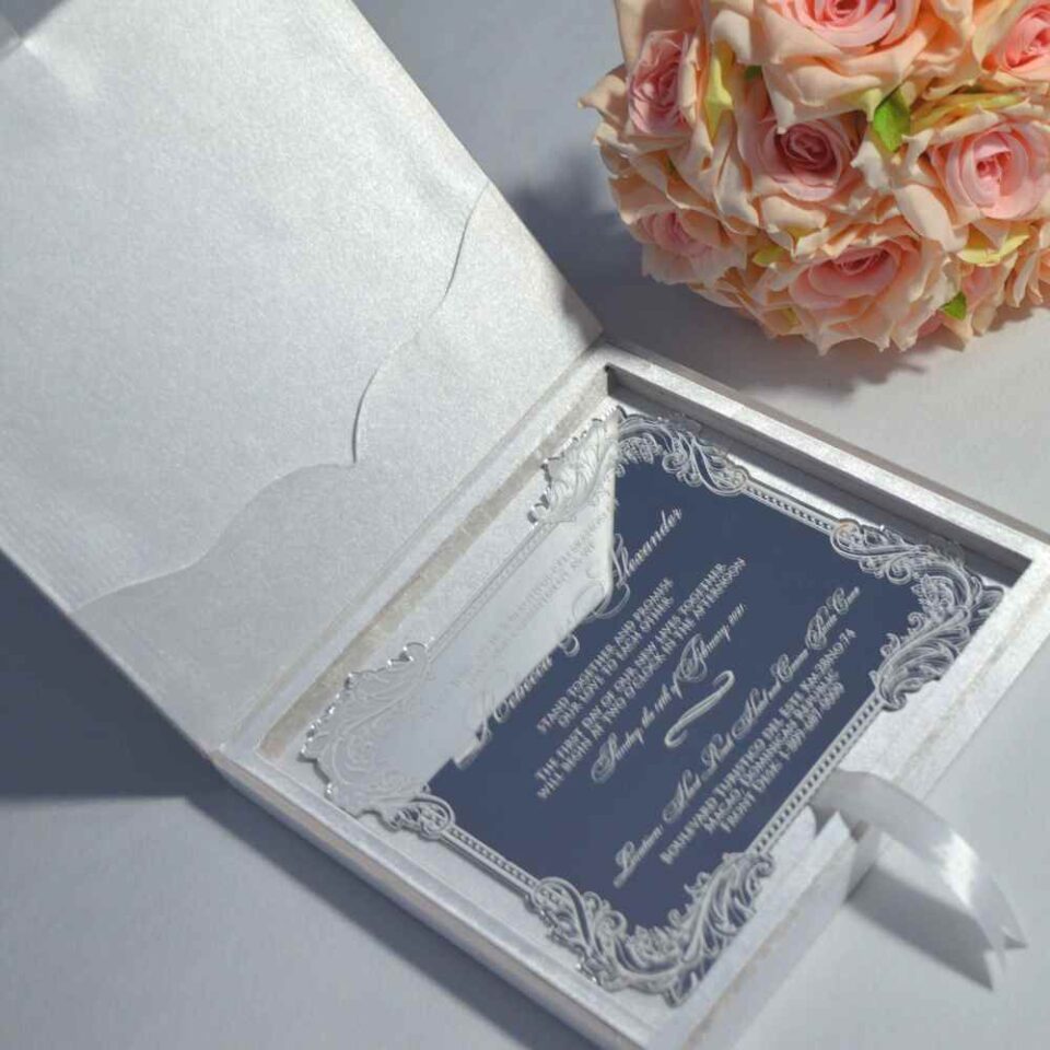 An open, white silk invitation box featuring a large, ornate gold monogram plaque on the left panel. Leaning against the right side is a clear acrylic wedding invitation with intricate silver-etched borders and white script lettering for the name. The invitation details a ceremony at the Hard Rock Hotel and Casino. In the background, soft peach-colored roses add a delicate floral accent against a neutral grey surface.