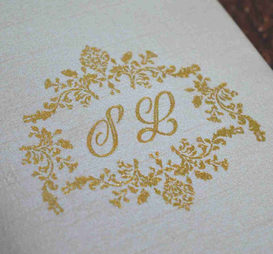 A high-angle, close-up shot of a **luxurious invitation box** with an ornate, three-dimensional lid design. The box features: * A **laser-cut gold wooden frame** on the lid with intricate floral and scrollwork patterns. * A central ** plaque** with a custom gold-etched monogram "R & F" in an elegant script. * A border of **sparkling clear crystals** embedded along the inner edge of the gold frame, adding a high-shine detail. * The rich, deep red velvet fabric covers the sides of the box, providing a soft, tactile contrast to the metallic gold top. * The box is displayed on a neutral, textured light-colored surface, highlighting its sophisticated and artisanal craftsmanship.