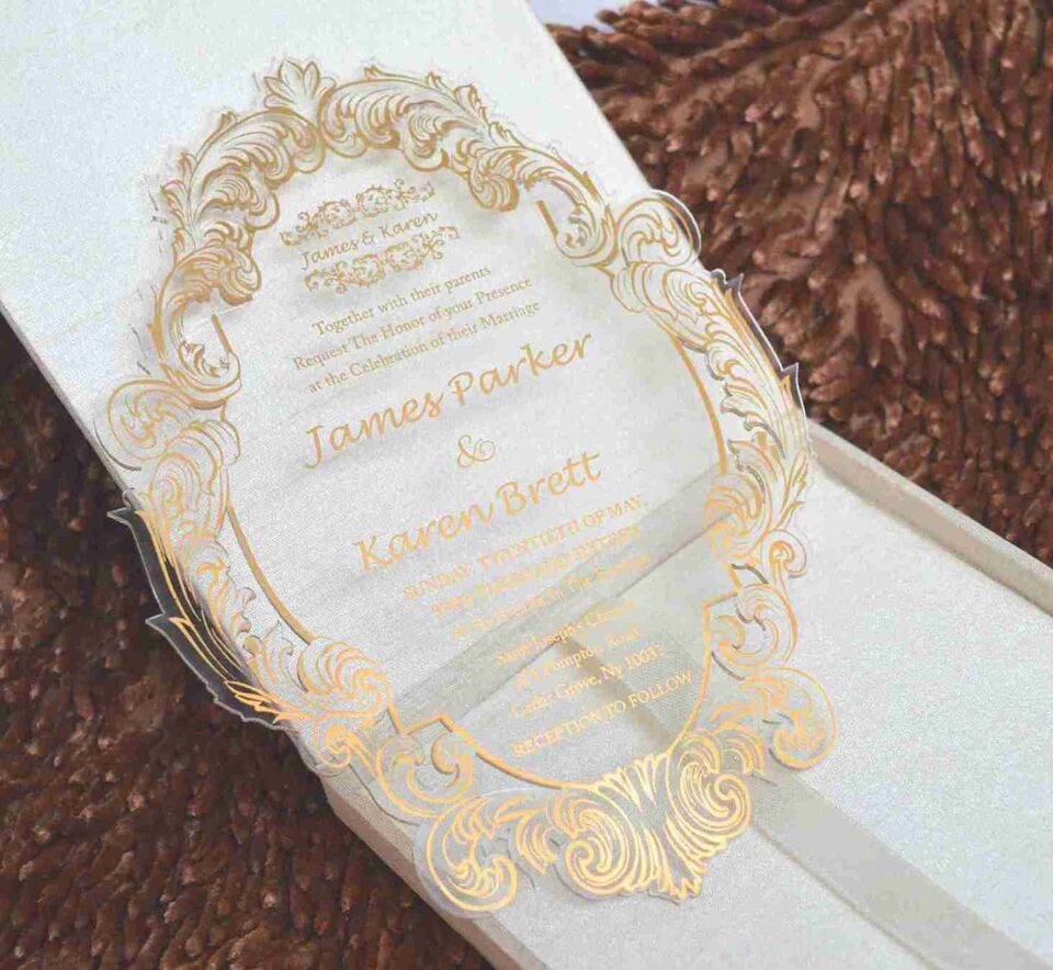 An open, **luxurious invitation box** shown from a high angle, revealing a coordinated stationery suite. The interior presentation includes: * **The Main Invitation:** A rectangular **gold clear acrylic card** featuring white etched calligraphy for the wedding of "Ronaldo and Fabiana." * **The Inner Lid:** A deep burgundy surface featuring a **gold-foiled custom monogram** and a romantic quote from Song of Solomon 3:4. * **Coordinated Inserts:** A matching burgundy paper card with gold-foiled event details is tucked beneath the acrylic invitation. * **Design Details:** The box is lined with rich burgundy velvet, and the gold-edged lid highlights the reflective quality of the acrylic card. * The entire set is displayed on a neutral, textured surface that emphasizes the deep jewel tones and metallic gold accents.