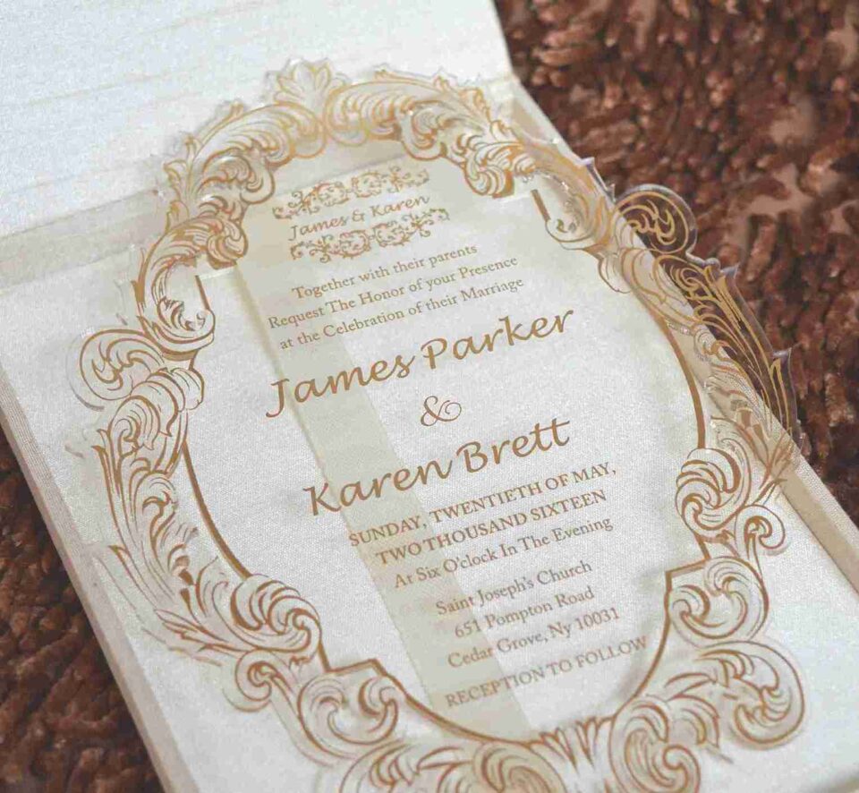 An open, **luxurious invitation box** shown from a high angle, revealing a coordinated stationery suite. The interior presentation includes: * **The Main Invitation:** A rectangular **gold clear acrylic card** featuring white etched calligraphy for the wedding of "Ronaldo and Fabiana." * **The Inner Lid:** A deep burgundy surface featuring a **gold-foiled custom monogram** and a romantic quote from Song of Solomon 3:4. * **Coordinated Inserts:** A matching burgundy paper card with gold-foiled event details is tucked beneath the acrylic invitation. * **Design Details:** The box is lined with rich burgundy velvet, and the gold-edged lid highlights the reflective quality of the acrylic card. * The entire set is displayed on a neutral, textured surface that emphasizes the deep jewel tones and metallic gold accents.