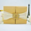 A front-facing view of a closed, square **champagne gold silk gatefold invitation folio** set against a white background. The luxury design features: * A wide **cream satin ribbon** wrapped horizontally around the center, gathered in the middle. * A large, ornate **gold-tone clasp** encrusted with sparkling crystals that joins the two front panels. * Decorative accents in the background, including a small, dark bronze **Eiffel Tower figurine** on the right and a soft white rose on the left. The shimmering gold silk and rhinestone-encrusted buckle create a sophisticated and glamorous presentation for a formal wedding or gala.