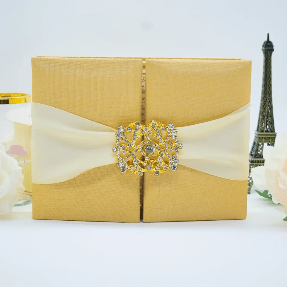 A front-facing view of a closed, square **champagne gold silk gatefold invitation folio** set against a white background. The luxury design features: * A wide **cream satin ribbon** wrapped horizontally around the center, gathered in the middle. * A large, ornate **gold-tone clasp** encrusted with sparkling crystals that joins the two front panels. * Decorative accents in the background, including a small, dark bronze **Eiffel Tower figurine** on the right and a soft white rose on the left. The shimmering gold silk and rhinestone-encrusted buckle create a sophisticated and glamorous presentation for a formal wedding or gala.