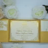 A high-angle view of an open **gold silk gatefold invitation folio** resting on a white surface, surrounded by white and peach-colored roses. The elegant wedding suite features: * **The center panel:** A formal cream-colored invitation card with a shimmering gold-foiled border and an ornate crest. It announces the marriage of "**Stephanie Elaine Godoy**" and "**William Benjamin Schilling**." The card is secured by matching gold silk corner tabs. * **Side pockets:** The left and right panels contain pockets holding coordinating stationery. On the left, two small cards with gold monograms and the word "Reception" are visible. * **Decorative accents:** A small, dark bronze **Eiffel Tower figurine** is partially visible in the upper right corner, adding a touch of travel-inspired romance. The combination of textured gold silk and metallic gold foil creates a regal and sophisticated aesthetic for a formal wedding celebration.