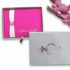 A rectangular, vibrant hot pink silk wedding invitation folio is centered against a white background. A wide, shimmering white or silver satin ribbon runs vertically down the left side, cinched at the middle by a round, sparkling rhinestone button brooch. In the bottom right corner, the names "Mathew and Michelle" are printed in an elegant, silver-toned cursive script. To the right of the folio, a tall stalk of bright pink gladiolus blossoms adds a soft, floral frame to the composition.