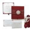 An open, luxury wedding invitation box in a deep burgundy red. The main invitation card is held in the base of the box with red corner tabs; it features the names "Oliver Edwards & Emily Luna" in an elegant script, surrounded by intricate silver or light grey filigree at the corners. The inner lid of the box also displays the couple's names and venue details on a matching white card. To the right of the box, decorative red rose petals and a string of white pearls are scattered against a white background. The box is angled, showing the depth of the red velvet-like interior.