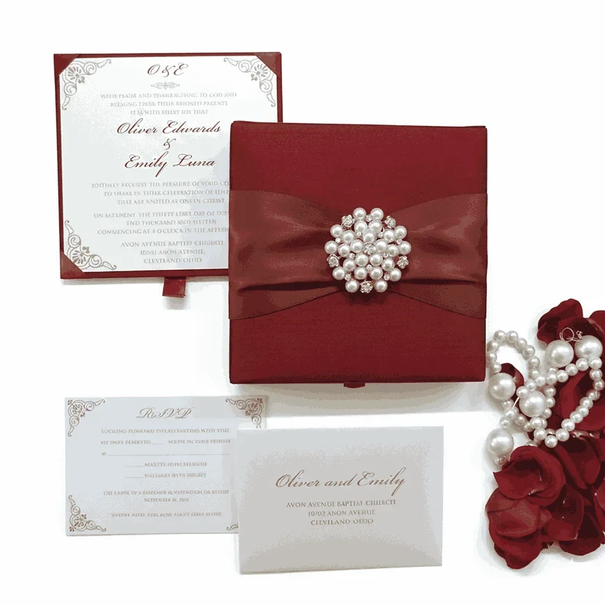 An open, luxury wedding invitation box in a deep burgundy red. The main invitation card is held in the base of the box with red corner tabs; it features the names "Oliver Edwards & Emily Luna" in an elegant script, surrounded by intricate silver or light grey filigree at the corners. The inner lid of the box also displays the couple's names and venue details on a matching white card. To the right of the box, decorative red rose petals and a string of white pearls are scattered against a white background. The box is angled, showing the depth of the red velvet-like interior.