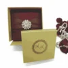 A square, deep red silk-textured invitation box centered on a white background. A wide, matching red satin ribbon wraps horizontally around the box, cinched in the middle by a large, circular pearl and crystal brooch. To the right of the box, a scattered pile of dark red rose petals is intertwined with a string of white pearls of varying sizes. The flat lay composition is brightly lit, highlighting the rich fabric texture and the contrast between the red elements and the clean white surface.