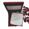 An open, luxury wedding invitation box in a deep burgundy red. The main invitation card is held in the base of the box with red corner tabs; it features the names "Oliver Edwards & Emily Luna" in an elegant script, surrounded by intricate silver or light grey filigree at the corners. The inner lid of the box also displays the couple's names and venue details on a matching white card. To the right of the box, decorative red rose petals and a string of white pearls are scattered against a white background. The box is angled, showing the depth of the red velvet-like interior.
