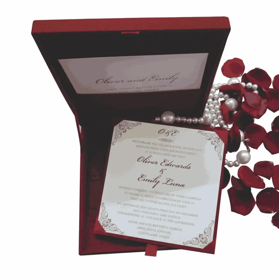 An open, luxury wedding invitation box in a deep burgundy red. The main invitation card is held in the base of the box with red corner tabs; it features the names "Oliver Edwards & Emily Luna" in an elegant script, surrounded by intricate silver or light grey filigree at the corners. The inner lid of the box also displays the couple's names and venue details on a matching white card. To the right of the box, decorative red rose petals and a string of white pearls are scattered against a white background. The box is angled, showing the depth of the red velvet-like interior.