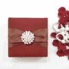 A square, deep red silk-textured invitation box centered on a white background. A wide, matching red satin ribbon wraps horizontally around the box, cinched in the middle by a large, circular pearl and crystal brooch. To the right of the box, a scattered pile of dark red rose petals is intertwined with a string of white pearls of varying sizes. The flat lay composition is brightly lit, highlighting the rich fabric texture and the contrast between the red elements and the clean white surface.