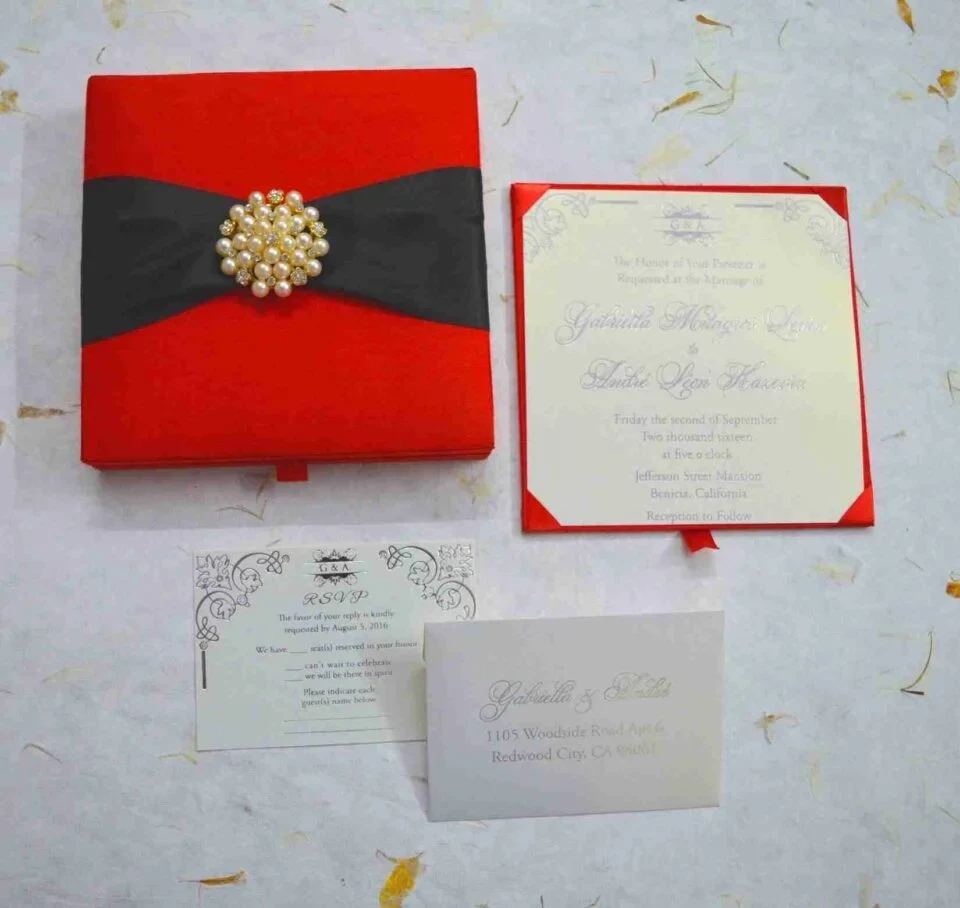 An open, square wedding invitation box in vibrant red silk. The main invitation card is nestled in the base, featuring the names "Gabriella Milagros Levin" and "André Leon Hazevra" in elegant grey script, topped with a small "G & A" monogram and decorative corner flourishes. The inner lid of the box contains a pocket holding a matching white card. The box is set on a white textured surface scattered with delicate yellow petals, creating a formal and colorful presentation.