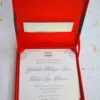 An open, square wedding invitation box in vibrant red silk. The main invitation card is nestled in the base, featuring the names "Gabriella Milagros Levin" and "André Leon Hazevra" in elegant grey script, topped with a small "G & A" monogram and decorative corner flourishes. The inner lid of the box contains a pocket holding a matching white card. The box is set on a white textured surface scattered with delicate yellow petals, creating a formal and colorful presentation.