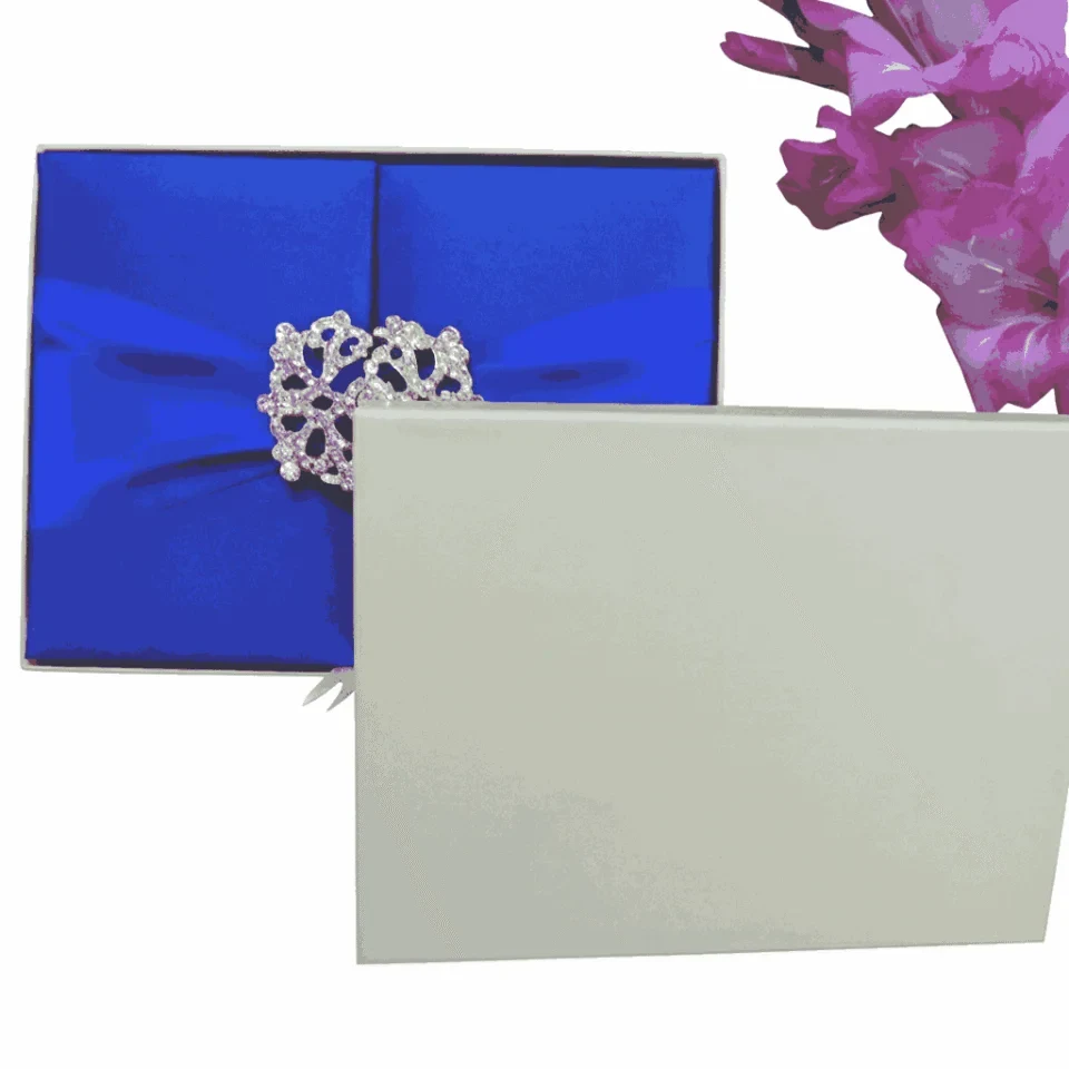 A rectangular, royal blue silk gatefold invitation box centered on a white background. A wide, matching blue satin ribbon wraps horizontally around the box, gathered in the center by a large, ornate silver brooch with a complex, lace-like filigree design. To the right of the box, a cluster of vibrant pink gladiolus flowers adds a pop of contrasting color. The flat lay composition is brightly lit, emphasizing the smooth sheen of the satin and the rich texture of the silk fabric.